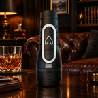 A black portable water bottle with a large white and black push-button design stands on a dark wooden table in a dimly lit, luxurious room with a brown leather sofa, bookshelves, and a glass of whiskey nearby.